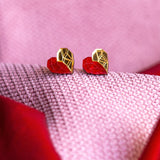 Gold and red geometric heart studs rest on pink and red textured fabric, with the colors and patterns creating a soft, romantic background—a perfect touch of romantic jewelry.
