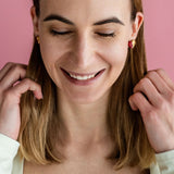 A woman with shoulder-length brown hair and a white top smiles with her eyes closed against a pink background, touching her hair with both hands, while wearing delicate handcrafted earrings.