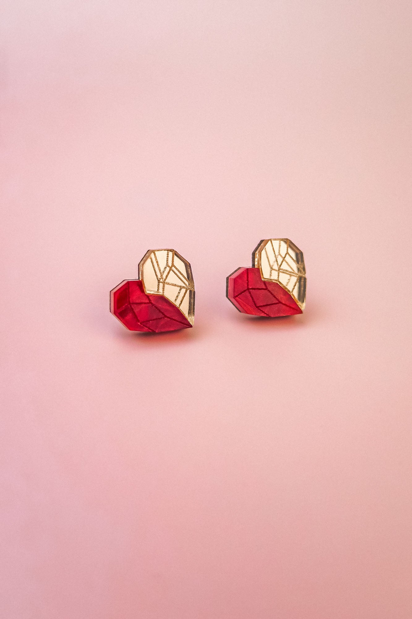 Two origami hearts, each half red and half gold, are placed side by side on a smooth, light pink background, resembling handcrafted earrings or romantic jewelry.