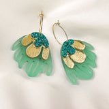 A pair of handcrafted earrings with gold metallic petals, glittery green accents, and translucent green acrylic shapes resembling leaves, displayed on white satin fabric—perfect for adding a touch of tropical elegance.