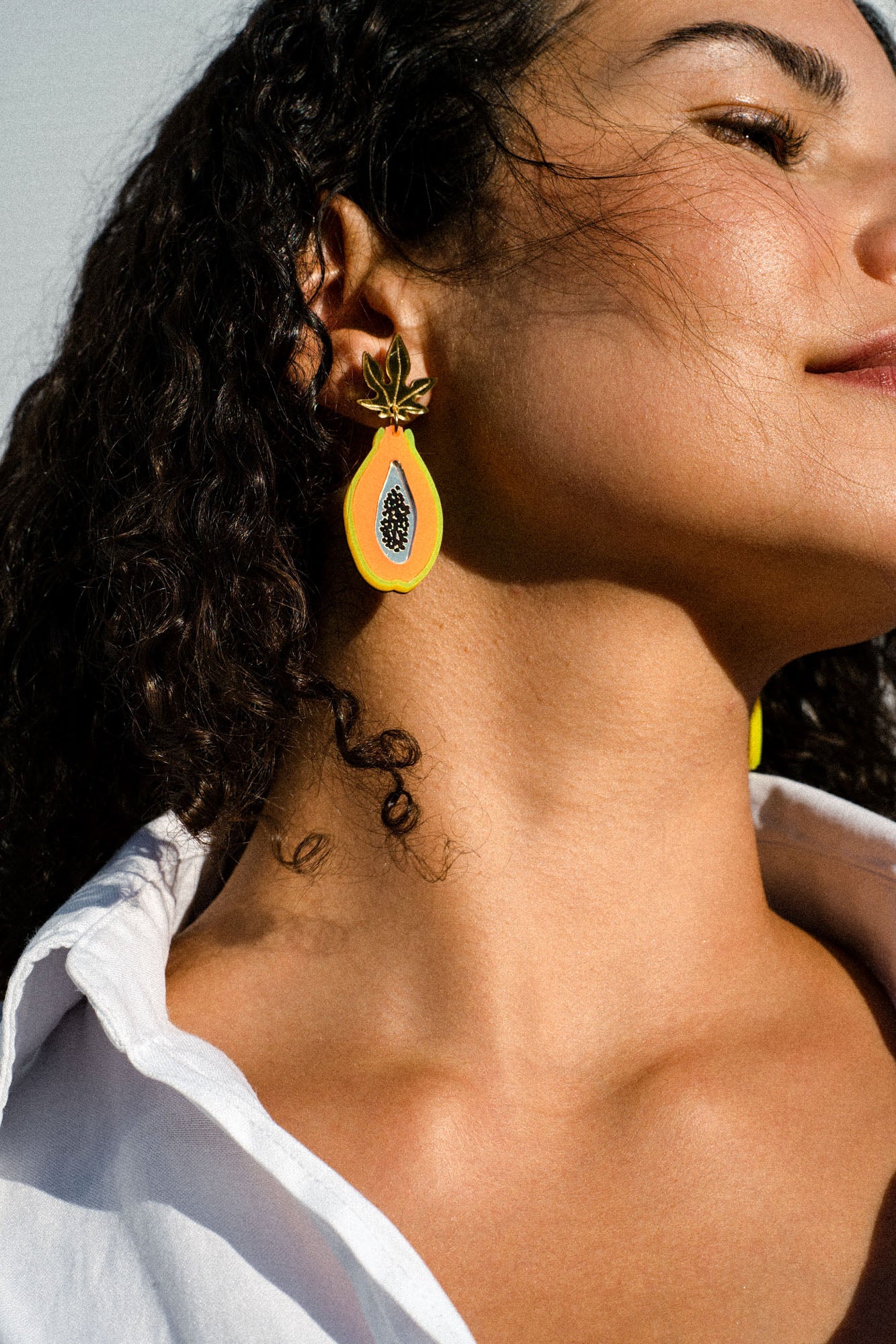 A woman with curly dark hair wears a white shirt and a colorful pair of Papaya Earrings, looking upward against a light background. These vibrant, handcrafted earrings add a playful touch to her modern style.