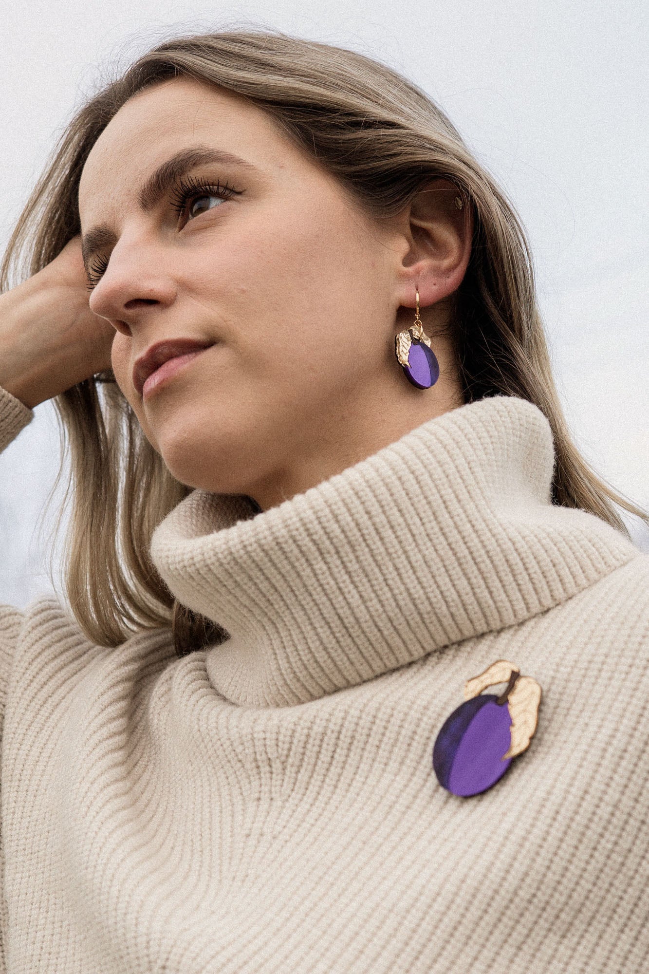 A woman with blonde hair wearing a cream-colored turtleneck sweater, handcrafted purple plum earrings, and a matching purple brooch stands outdoors, looking thoughtful with her hand resting on her head.
