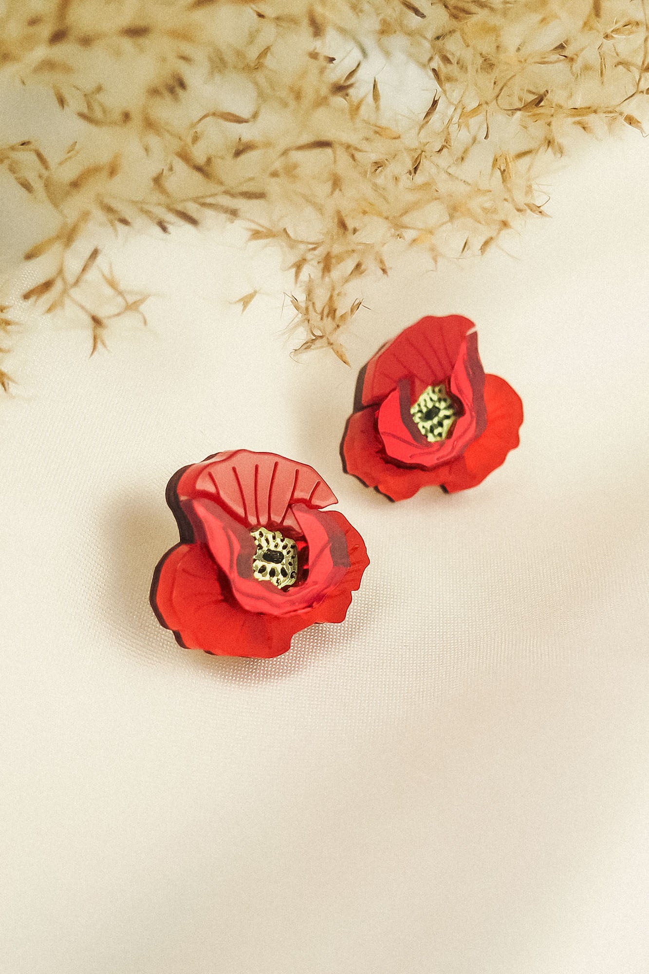 Two red poppy design acrylic earrings with black and yellow centers rest on a white fabric surface. Beige dried foliage in the background creates a soft, elegant setting, highlighting their unique style and lightweight feel.