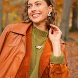 A woman smiles elegantly while standing in a forest with autumn leaves. She wears a brown leather coat over an orange suede jacket, with a green sweater, patterned skirt, and a handcrafted Tulip Necklace. Tall trees and fallen leaves fill the background.