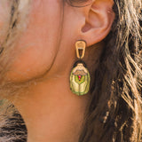 A close-up of a womans ear adorned with Rose Chafer Earrings, handcrafted and nature-inspired, featuring an illustrated figure in green clothing. Her brown hair partially covers her ear while dried grass adds texture to the foreground.