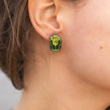 Close-up of a person wearing Rose Chafer Studs—nature-inspired earrings with green gemstones and gold details. The person has wavy brown hair, a green top, and is set against a softly blurred background.