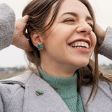 A woman with straight, light brown hair and light skin wears a light pink top and turquoise Sea Turtle Studs, standing in front of a mauve curtain, gazing slightly to the left for a summery vibe.