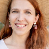 A woman with long auburn hair wearing a wide-brimmed straw hat, lightweight seashell earrings, and a light V-neck top smiles outdoors with greenery in the background.