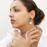 A woman with brown hair in a low bun wears a white blazer, gold rings, and lightweight seashell earrings with turquoise accents. She sits with her hands gently touching her neck, gazing thoughtfully to the side.
