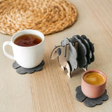A white mug and a pink cup sit on dark felt coasters next to a decorative wooden sheep-shaped coaster holder, bringing charming home decor touches to a light wooden table topped with a round woven placemat.