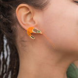 A person with brown skin and dreadlocks wears a detailed, neon orange Shrimp Stud earring adorned with emerald rhinestones. Their hand rests on their head, and they are dressed in a green top.