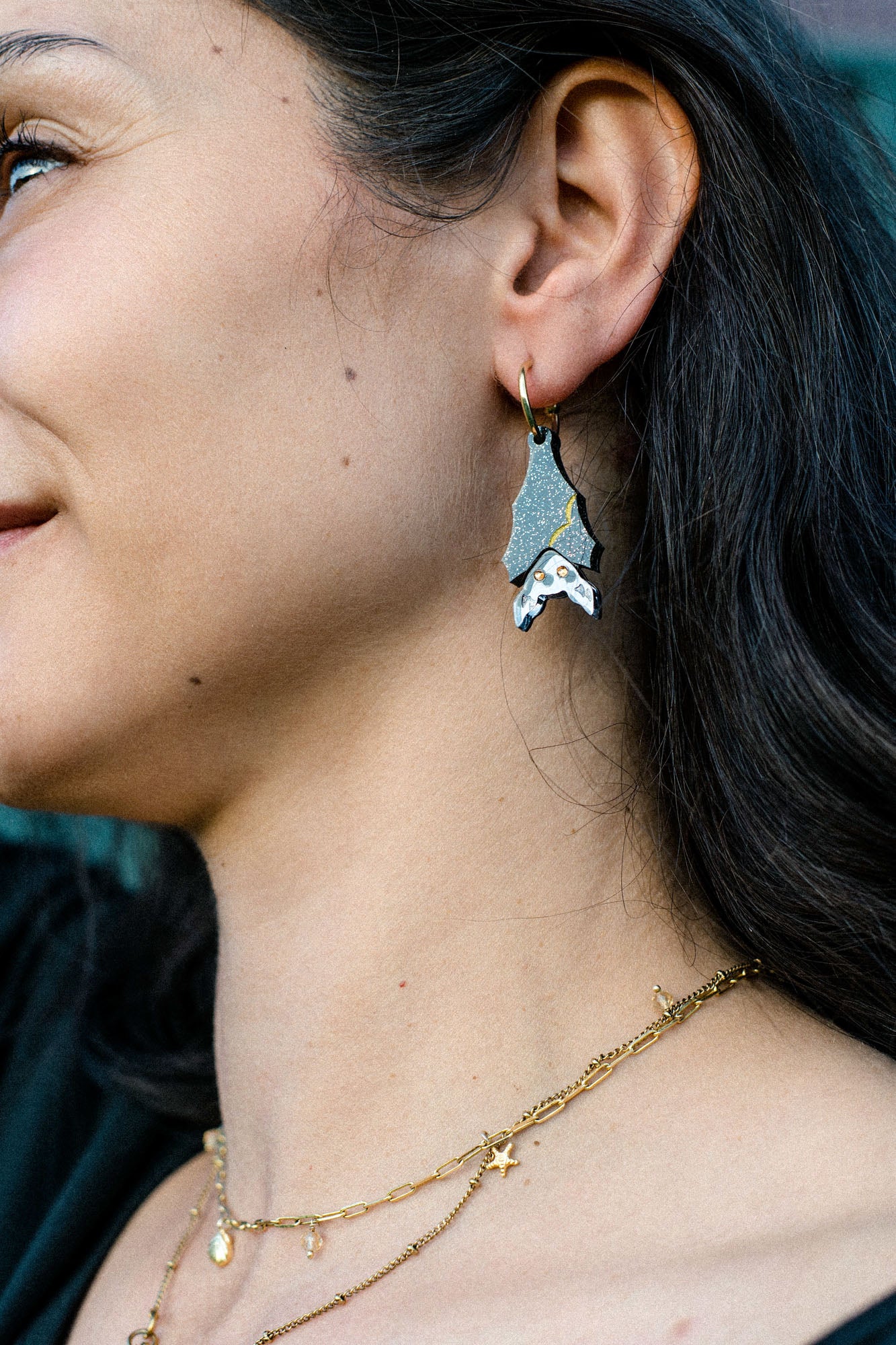 A close-up of a woman's face and neck, showing her wearing a gothic glam Spooky Bat earring in silver and black, paired with layered gold necklaces with small charms. She has long dark hair and a slight smile.