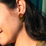 Close-up of a person with dark hair wearing striking triangle earrings crafted from acrylic glass, paired with a gold chain necklace. The background features blurred green vertical lines, adding to the modern aesthetic.