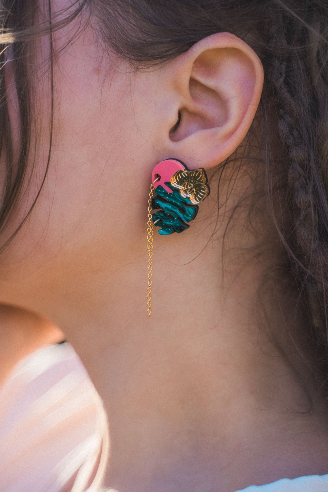 Close-up of a person’s ear wearing Tropicana Earrings—a tropical-inspired design with a gold tiger head, blue and pink details, and a delicate gold chain. The person’s hair is partially braided.