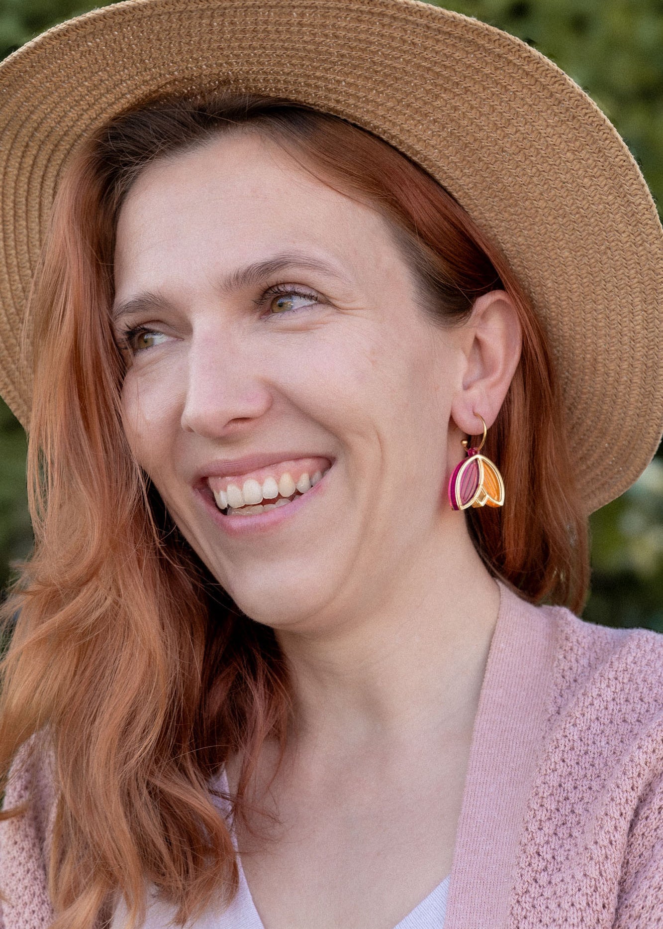 A woman with wavy reddish hair, wearing a straw hat, pink cardigan, white top, and Tulip Huggie Earrings featuring vibrant colors, smiles while standing outdoors in front of green foliage.