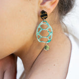 A close-up of a person’s ear wearing a statement, sustainable earring with a black geometric stud, a large mint green oval with cutouts, and a small gold disc at the bottom—showcasing zero-waste beauty. The person has light skin and dark brown hair.