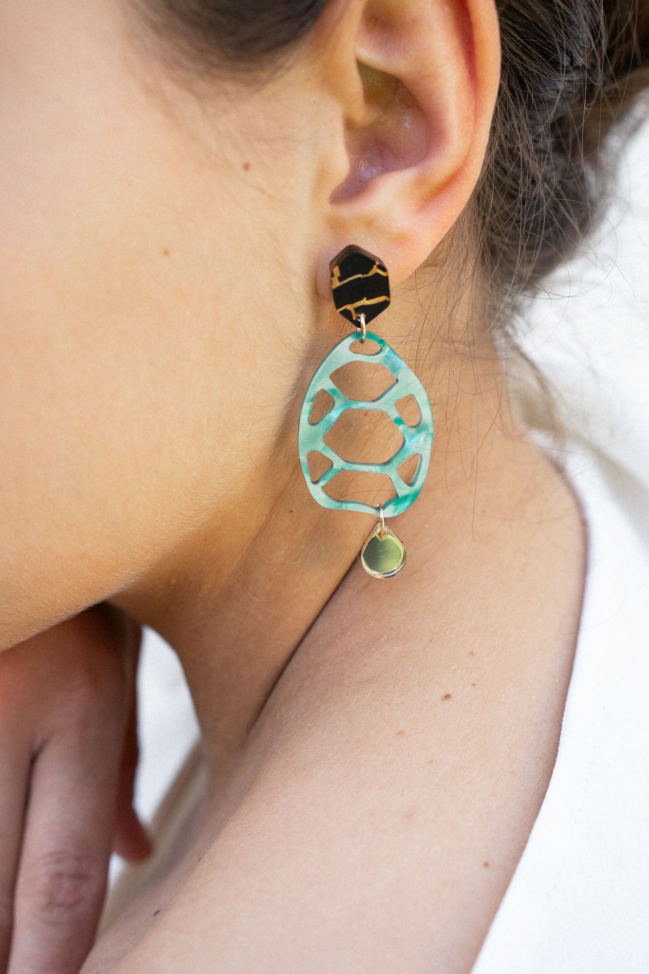 A close-up of a person’s ear wearing a statement, sustainable earring with a black geometric stud, a large mint green oval with cutouts, and a small gold disc at the bottom—showcasing zero-waste beauty. The person has light skin and dark brown hair.