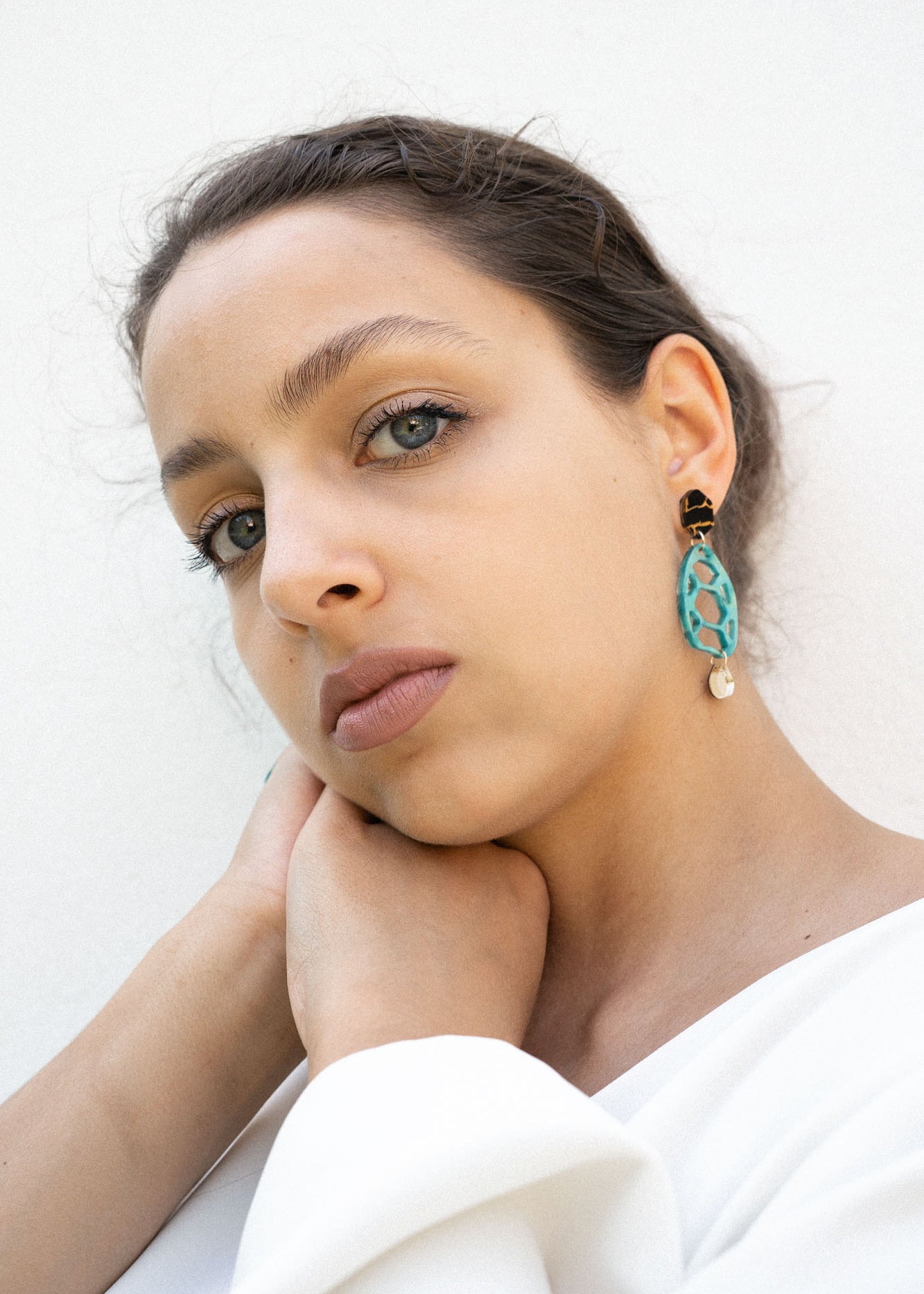 A woman with light eyes and brown hair, wearing a white top, rests her chin on her hand. She wears sustainable turquoise and gold earrings and looks calmly into the camera against a plain white background.