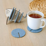 A white ribbed ceramic mug sits on a round gray felt coaster on a wooden table, with Whale Coasters and a whale-shaped holder nearby. A woven placemat adds home charm while these coasters help protect surfaces in style.