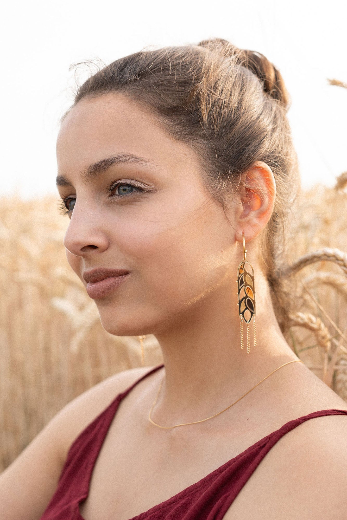 A woman with light brown hair in a bun, wearing a sleeveless burgundy top, gold necklace, and Wheat Statement Earrings, sits in a wheat field, looking thoughtfully to the side—her standout style capturing quiet elegance.