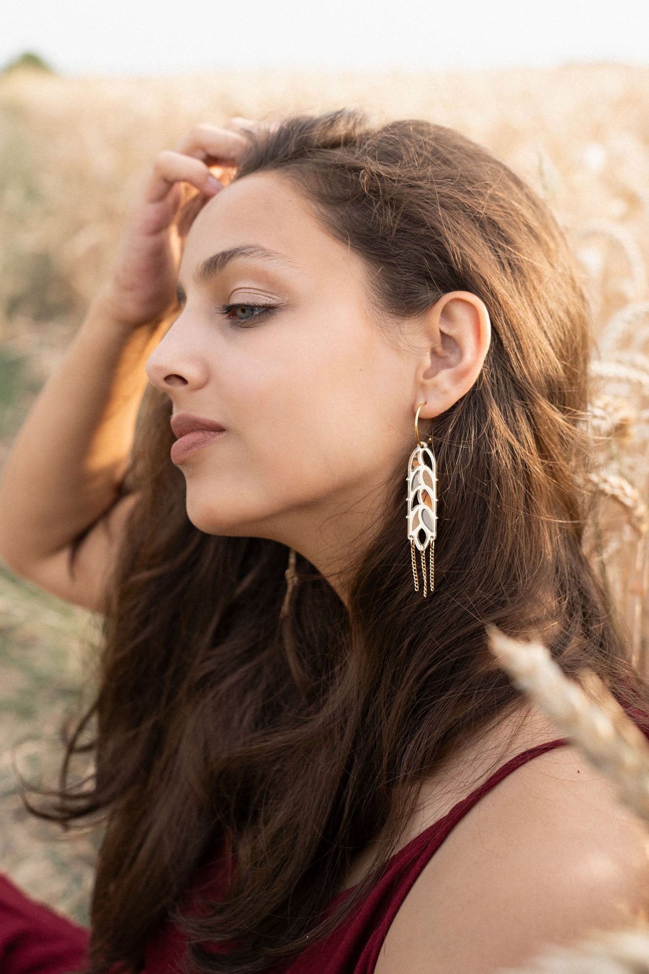 A young woman with long brown hair sits in a wheat field, wearing a burgundy dress and Wheat Statement Earrings. She gazes thoughtfully to the side, lightly touching her hair, her standout style enhanced by the bold design of her earrings.