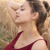 A young woman in a burgundy dress sits in a wheat field with her eyes closed, one arm raised behind her head. She wears Wheat Statement Earrings and a delicate necklace, appearing relaxed and peaceful in the natural setting with standout style.