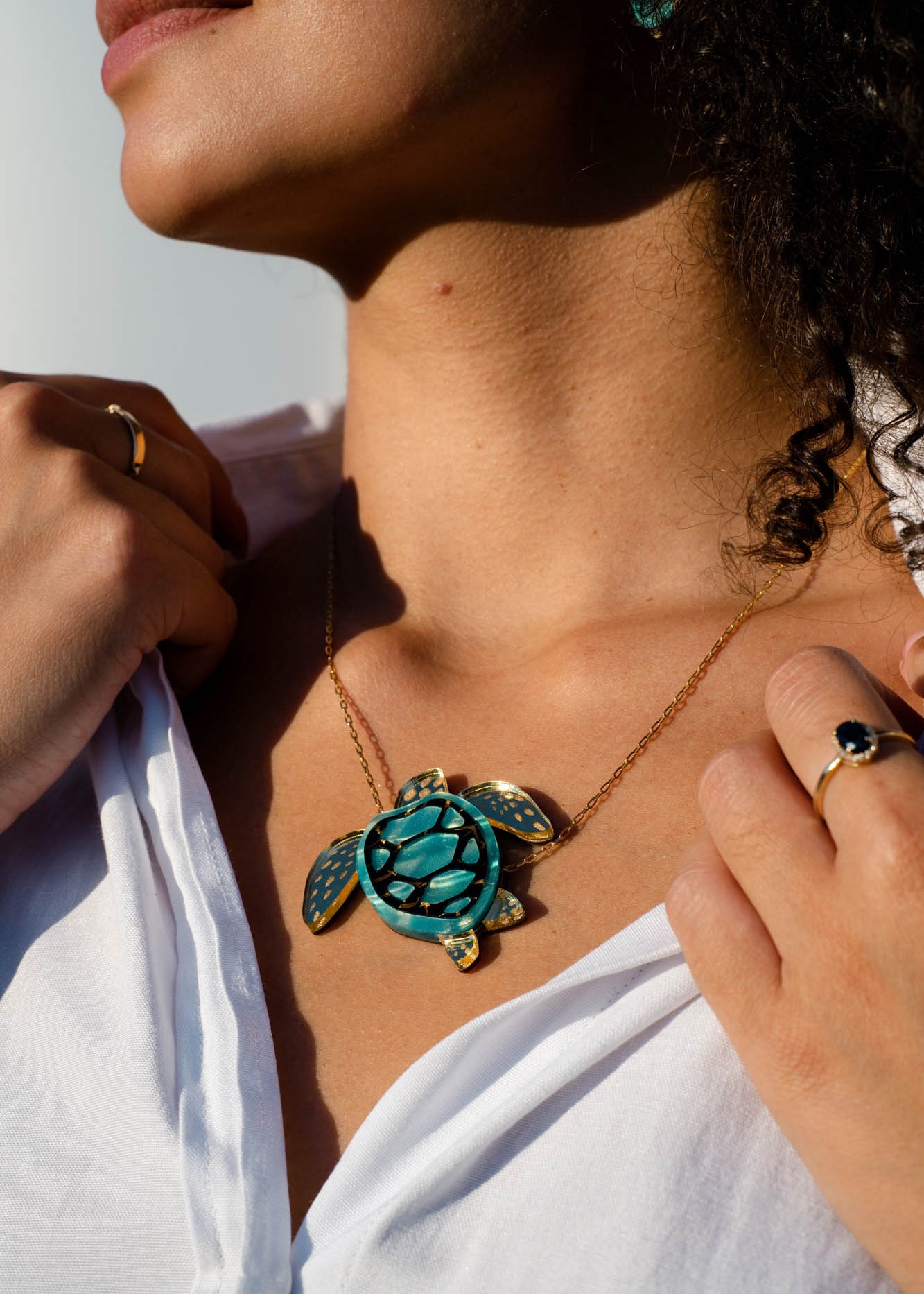A woman wearing a white shirt displays a handcrafted Sea Turtle Necklace featuring a gold chain and a turquoise turtle pendant. She holds her shirt collar with both hands, showing rings on her fingers. Sunlight highlights her jewelry and skin.