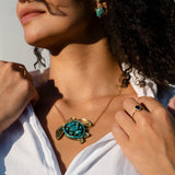 A woman in a white shirt wears a stunning turquoise and gold Sea Turtle Necklace, complemented by turquoise earrings and rings. Sunlight highlights her face and curly hair as she looks upward, casting a shadow across her cheek—a perfect moment to showcase her love for handcrafted jewelry.